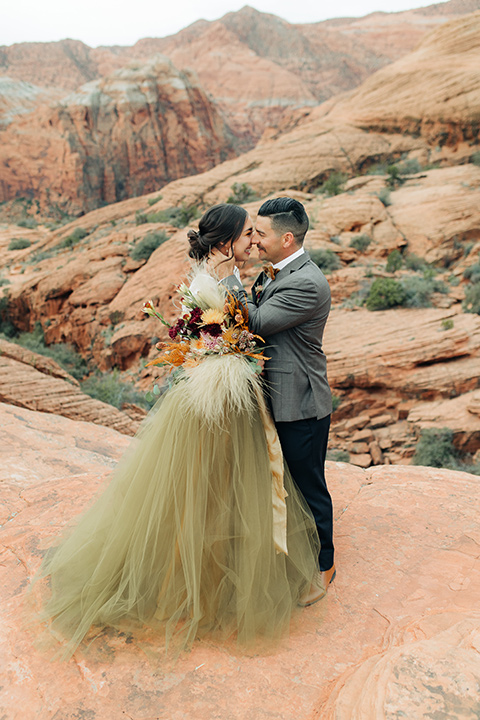 Utah-snow-canyon-shoot-bride-and-groom-laughing-and-touching-heads-the-bride-is-wearing-an-olive-green-tulle-skirt-and-an-ivory-lace-crop-top-with-long-sleeves-the-groom-in-a-café-brown-suit-with-blue-navy-pants-and-vest-with-a-gold-velvet-bow-tie