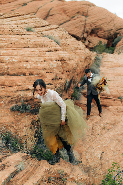 Utah-snow-canyon-shoot-bride-and-groom-with-boots-hiking-the-bride-is-wearing-an-olive-green-tulle-skirt-and-an-ivory-lace-crop-top-with-long-sleeves-the-groom-in-a-café-brown-suit-with-blue-navy-pants-and-vest-with-a-gold-velvet-bow-tie