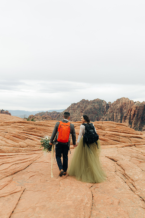 Utah-snow-canyon-shoot-bride-and-groom-with-hiking-packs-walking-away-the-bride-is-wearing-an-olive-green-tulle-skirt-and-an-ivory-lace-crop-top-with-long-sleeves-the-groom-in-a-café-brown-suit-with-blue-navy-pants-and-vest-with-a-gold-velvet-bow-tie