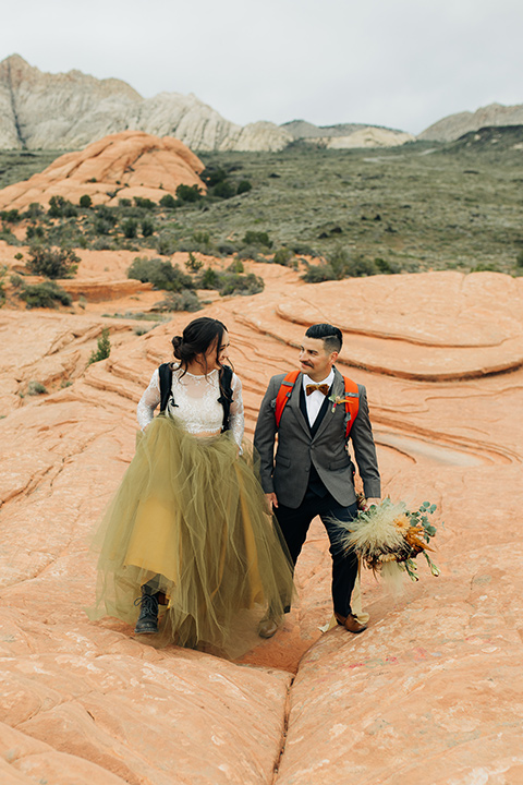 Utah-snow-canyon-shoot-bride-and-groom-with-hiking-packs-walking-towards-camera-bride-wearing-a-olive-green-tulle-skirt-with-an-ivory-lace-long-sleeve-crop-top-and-the-groom-is-in-a-café-brown-coat-with-a-blue-pair-of-pants-and-a-vest-with-a-gold-velvet-bow-tie