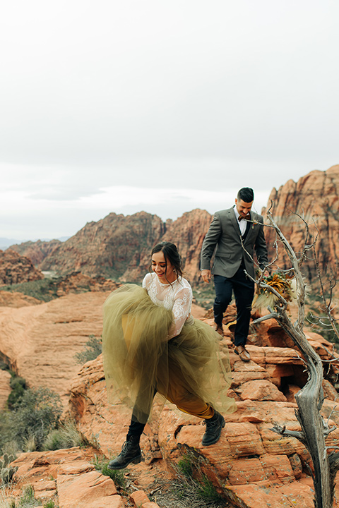 Utah-snow-canyon-shoot-bride-jumping-from-rock-to-rock-bride-wearing-a-olive-green-tulle-skirt-with-an-ivory-lace-long-sleeve-crop-top-and-the-groom-is-in-a-café-brown-coat-with-a-blue-pair-of-pants-and-a-vest-with-a-gold-velvet-bow-tie