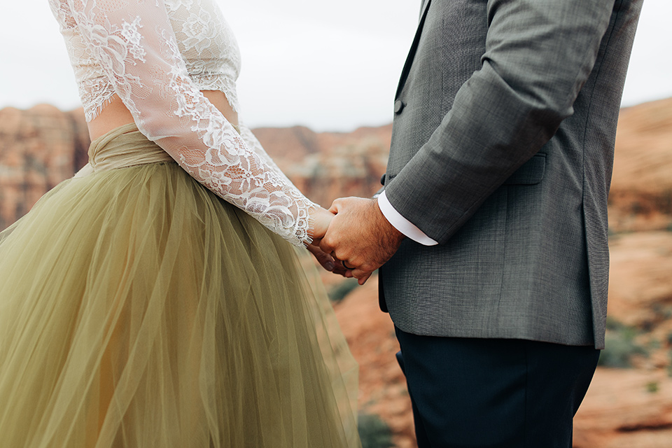 Utah-anow-canyon-shoot-close-up-of-bride-and-groom-holding-hands-the-bride-wears-an-olive-green-tulle-skirt-with-an-ivory-lace-long-sleeved-top-and-the-groom-in-a-café-brown-coat-with-a-dark-blue-pant-and-vest-with-a-gold-velvet-bow-tie