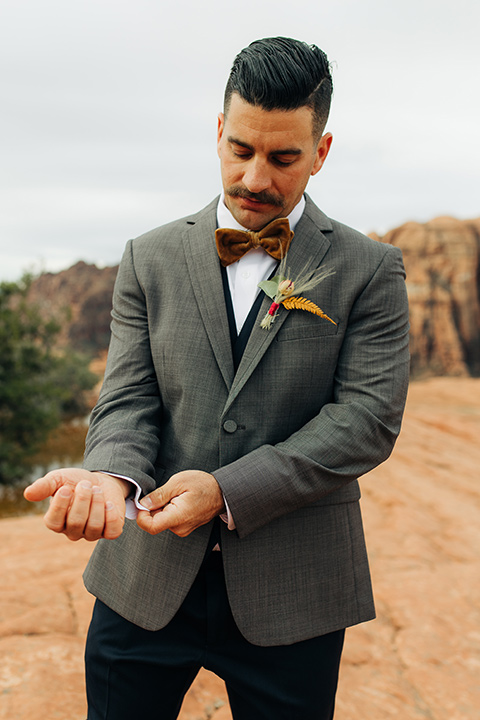 Utah-snow-canyon-shoot-groom-fixing-cuff-in-a-café-brown-coat-with-a-blue-pair-of-pants-and-a-vest-with-a-gold-velvet-bow-tie