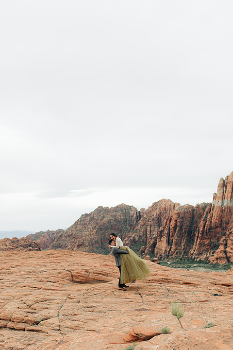 Utah-snow-canyon-shoot-groom-picking-up-bride-bride-wearing-a-olive-green-tulle-skirt-with-an-ivory-lace-long-sleeve-crop-top-and-the-groom-is-in-a-café-brown-coat-with-a-blue-pair-of-pants-and-a-vest-with-a-gold-velvet-bow-tie