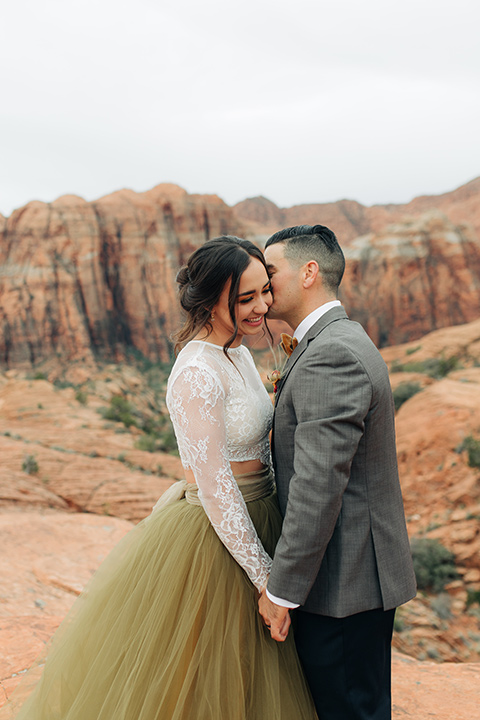 Utah-snow-canyon-shoot-groom-whispering-in-brides-ear-bride-wearing-a-olive-green-tulle-skirt-with-an-ivory-lace-long-sleeve-crop-top-and-the-groom-is-in-a-café-brown-coat-with-a-blue-pair-of-pants-and-a-vest-with-a-gold-velvet-bow-tie