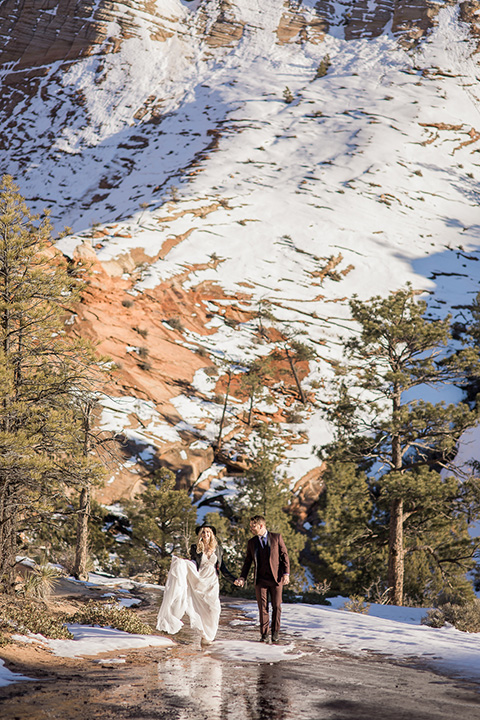 Utah-Shoot-bride-and-groom-crossing-stream-bride-in-a-tulle-flowing-gown-with-a-leather-jacket-and-the-groom-is-in-a-burgundy-tuxedo-with-a-black-long-tie