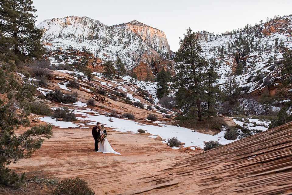 Utah-Shoot-bride-and-groom-far-away-shot-on-cliff-bride-in-a-flowing-tulle-gown-and-lace-bodice-with-a-leather-jacket-while-the-groom-is-in-a-burgundy-tuxedo-with-a-long-black-tie