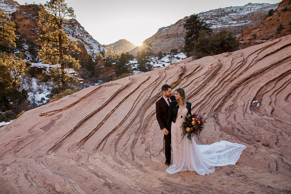 Utah-Shoot-bride-and-groom-standing-on-rock-cliff-bride-in-a-flowing-tulle-gown-and-lace-bodice-with-a-leather-jacket-while-the-groom-is-in-a-burgundy-tuxedo-with-a-long-black-tie