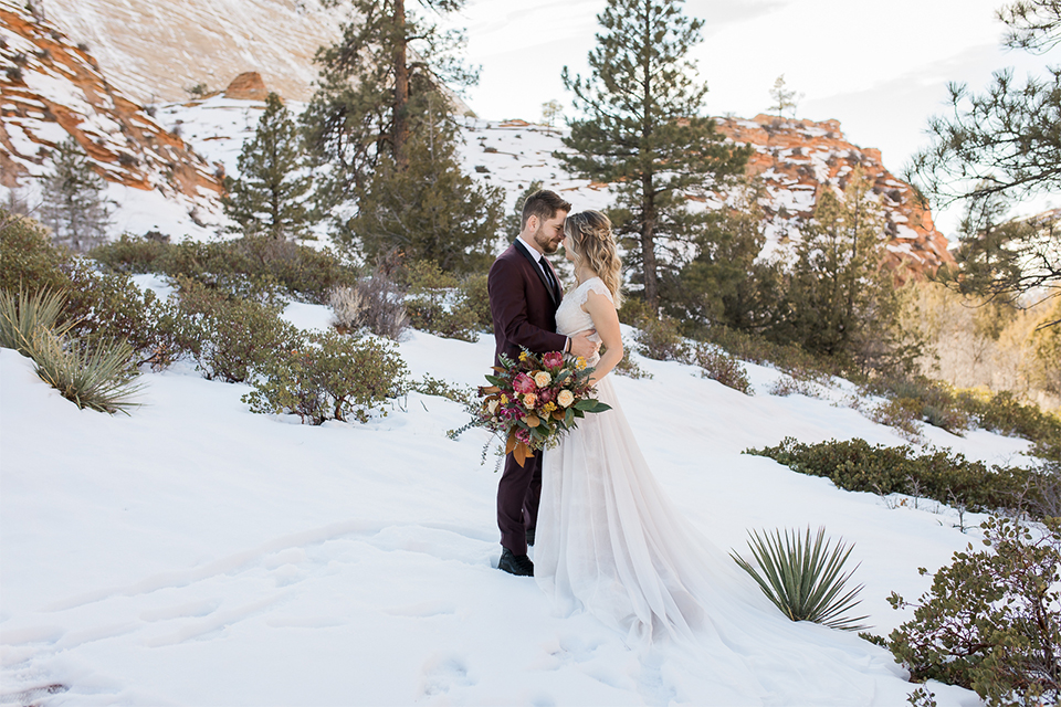 Utah-Shoot-bride-and-groom-touching-heads-bride-in-a-flowing-tulle-gown-and-lace-bodice-with-a-leather-jacket-while-the-groom-is-in-a-burgundy-tuxedo-with-a-long-black-tie