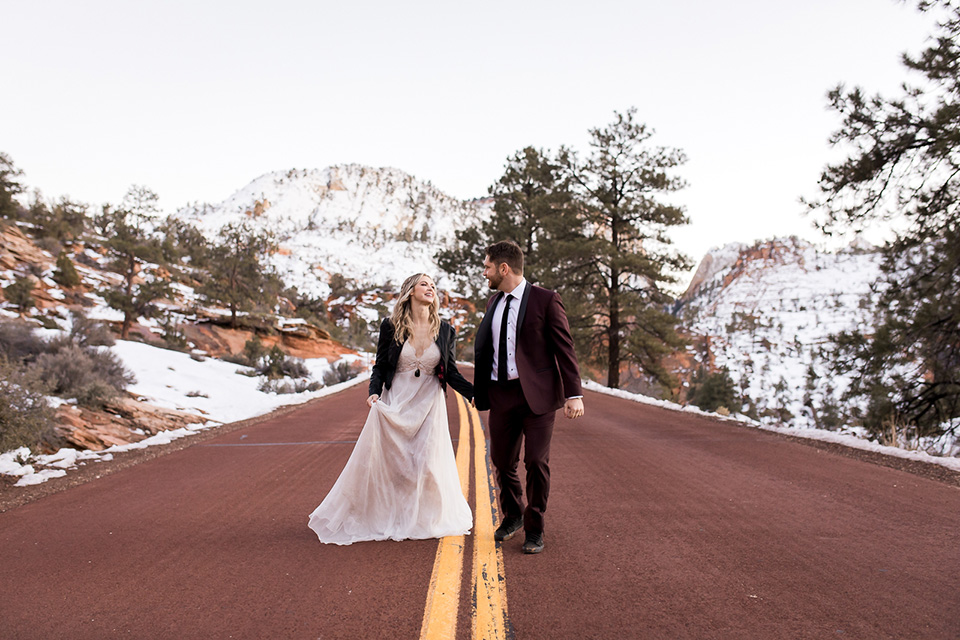 Utah-Shoot-bride-and-groom-walkking-on-the-road-bride-in-a-flowing-tulle-gown-and-lace-bodice-with-a-leather-jacket-while-the-groom-is-in-a-burgundy-tuxedo-with-a-long-black-tie