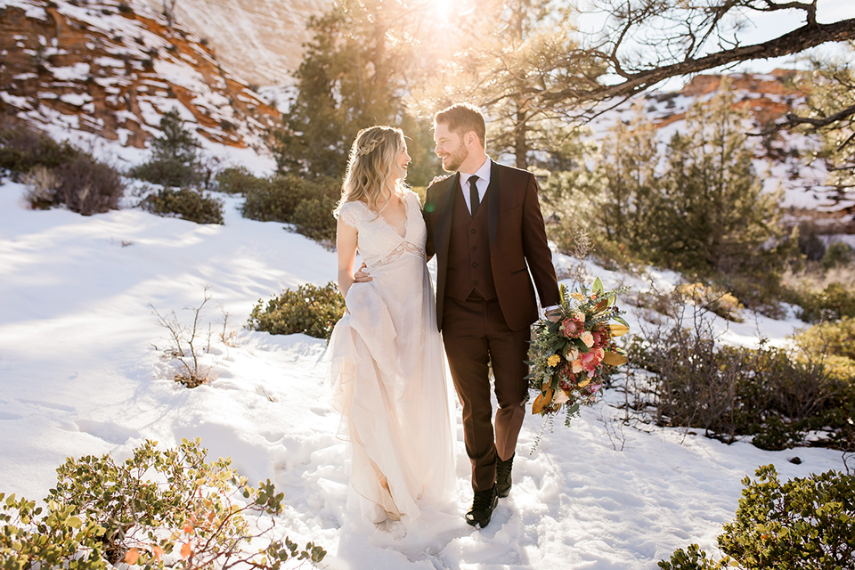 Utah-Shoot-couple-walking-in-snow-bride-in-a-flowing-tulle-gown-and-lace-bodice-with-a-leather-jacket-while-the-groom-is-in-a-burgundy-tuxedo-with-a-long-black-tie