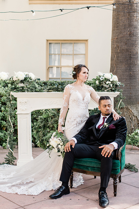 Maxwell-House-goom-sitting-in-a-chair-bride-standing-behind-them-bride-in-a-modern-lace-gown-groom-in-velvet-black-tuxedo