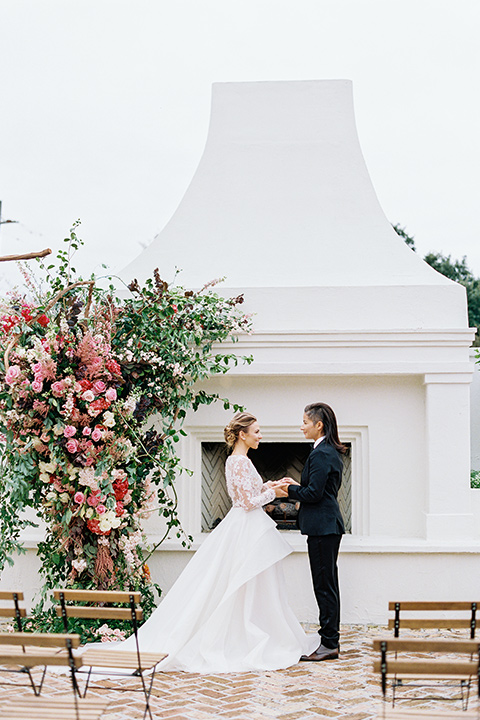 flower-market-shoot-brides-at-ceremony-space-bride-in-a-womens-black-shawl-lapel-tuxedo-with-polka-dot-bow-tie-bride-in-a-flowing-tulle-ball-gown-with-lace-sleeves-and-hair-up