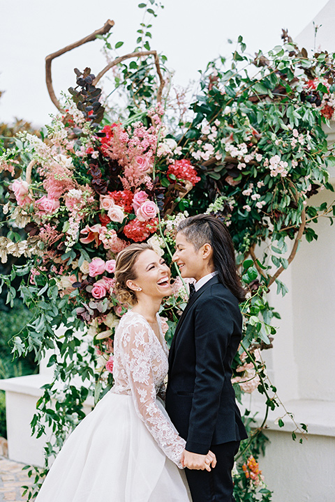 flower-market-shoot-brides-laughing-at-ceremony-bride-in-a-womens-black-shawl-lapel-tuxedo-with-polka-dot-bow-tie-bride-in-a-flowing-tulle-ball-gown-with-lace-sleeves-and-hair-up