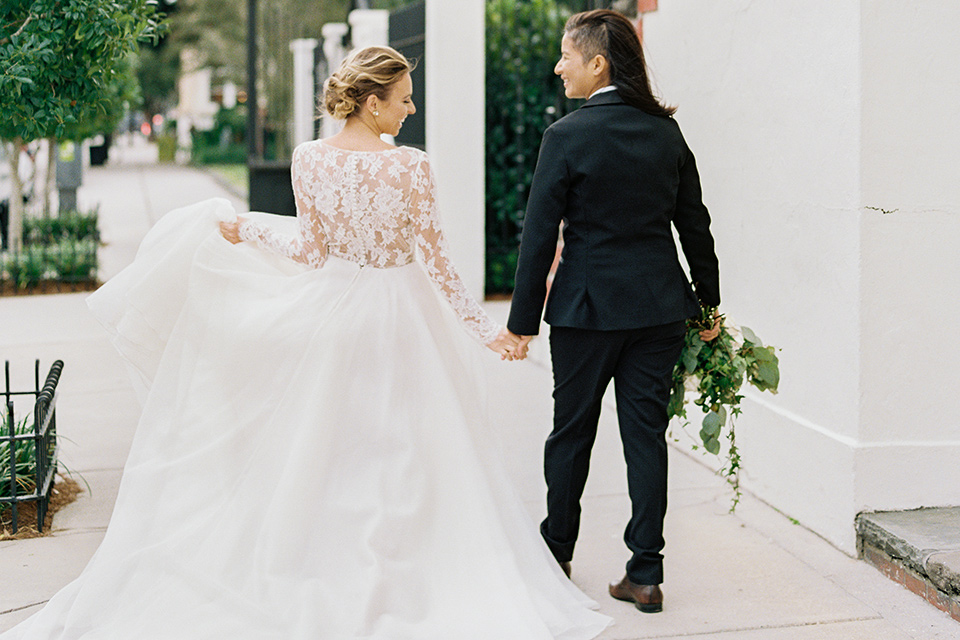 flower-market-shoot-brides-walking-away-from-camera-one-bride-in-a-flowing-tulle-gown-with-lace-sleeves-one-bride-in-a-blacl-shawl-lapel-tuxedo-with-a-polka-dot-bow