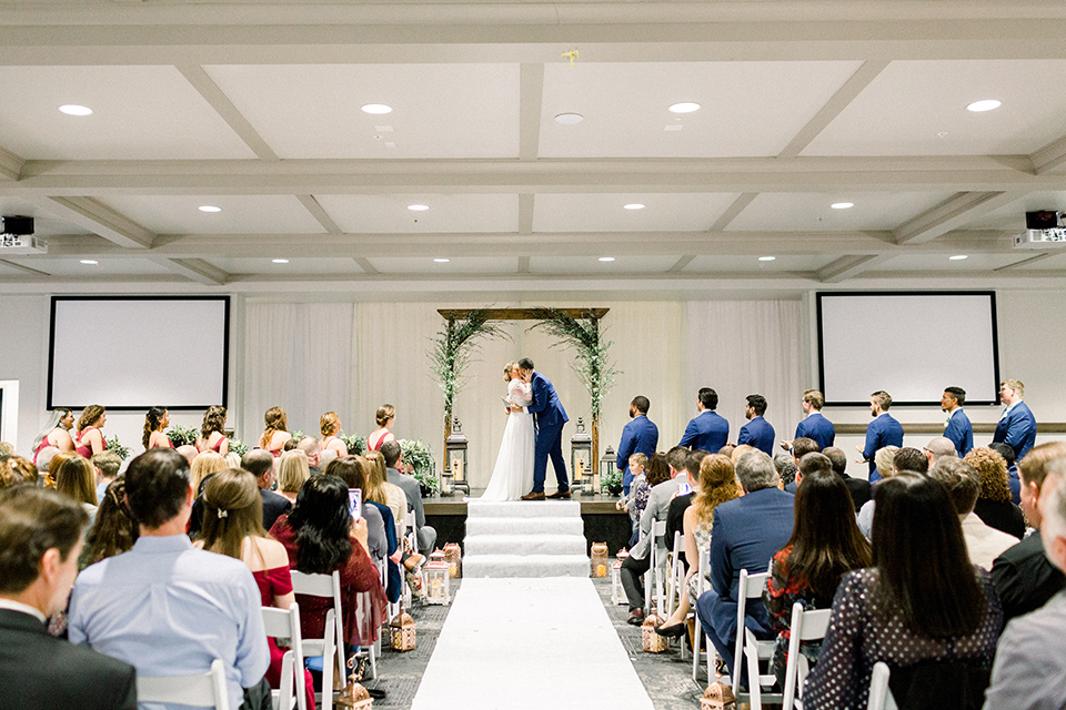 community-church-wedding-first-kiss-bride-in-a-flowing-gown-with-long-sleeves-and-an-illusion-bodice-groom-in-a-cobalt-blue-suit-with-a-berry-colored-tie