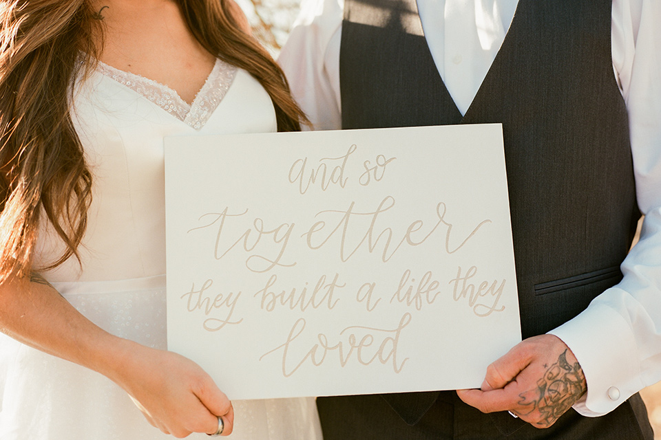 red-rocks-shoot-couple-holding-sign-bride-wearing-a-flowing-white-gown-and-groom-in-a-charcoal-grey-suit