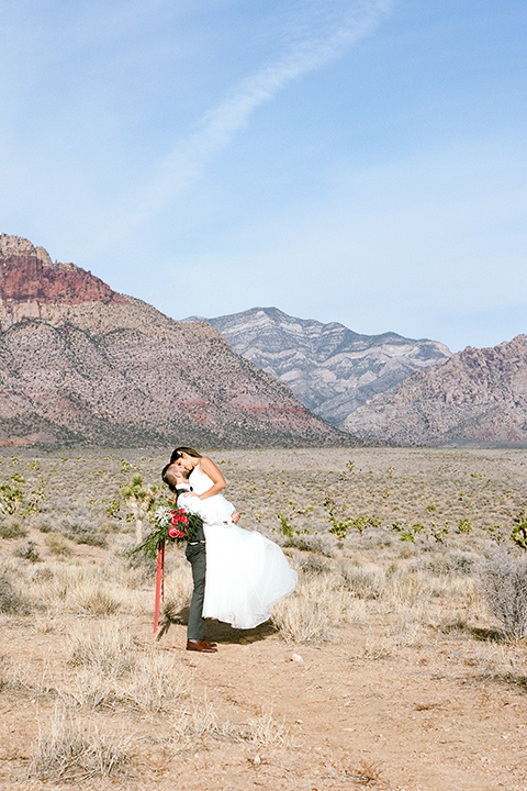 red-rocks-shoot-groom-holding-up-bride-bride-in-a-flowing-white-gown-groom-in-a-grey-suit