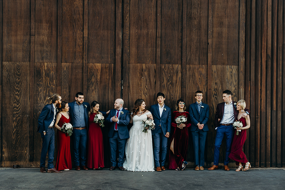 la-jolla-wedding-bridal-party-smiling-by-wooden-barn-bridesmaids-in-deep-red-dresses-groomsmen-in-dark-blue-suits-bride-in-a-flowing-ball-gown-with-a-detailed-bodice-and-straps-groom-in-a-dark-blue-suit-with-a-light-blue-suit-and-red-tie