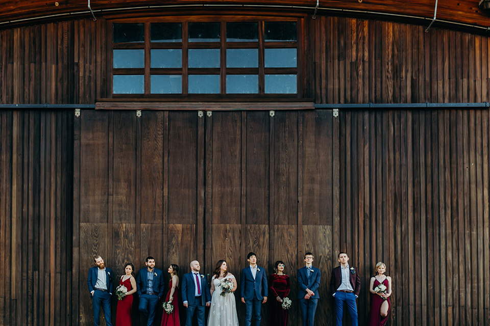 la-jolla-wedding-bridal-party-waist-up-photo-by-barn-bridesmaids-in-deep-red-dresses-groomsmen-in-dark-blue-suits-bride-in-a-flowing-ball-gown-with-a-detailed-bodice-and-straps-groom-in-a-dark-blue-suit-with-a-light-blue-suit-and-red-tie