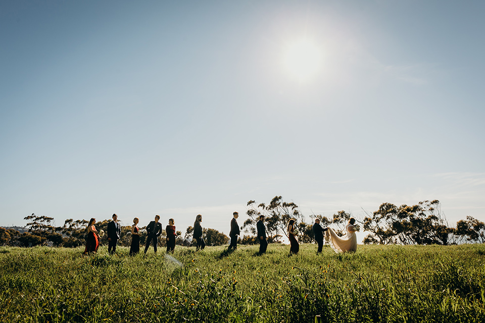 la-jolla-wedding-bridal-party-walking-in-a-line-bridesmaids-in-deep-red-dresses-groomsmen-in-dark-blue-suits-bride-in-a-flowing-ball-gown-with-a-detailed-bodice-and-straps-groom-in-a-dark-blue-suit-with-a-light-blue-suit-and-red-tie