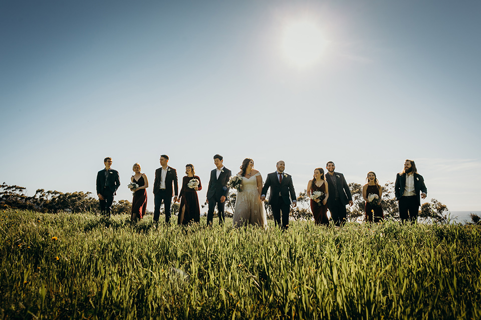 la-jolla-wedding-bridal-party-walking-in-grass-in-deep-red-dresses-groomsmen-in-dark-blue-suits-bride-in-a-flowing-ball-gown-with-a-detailed-bodice-and-straps-groom-in-a-dark-blue-suit-with-a-light-blue-suit-and-red-tie