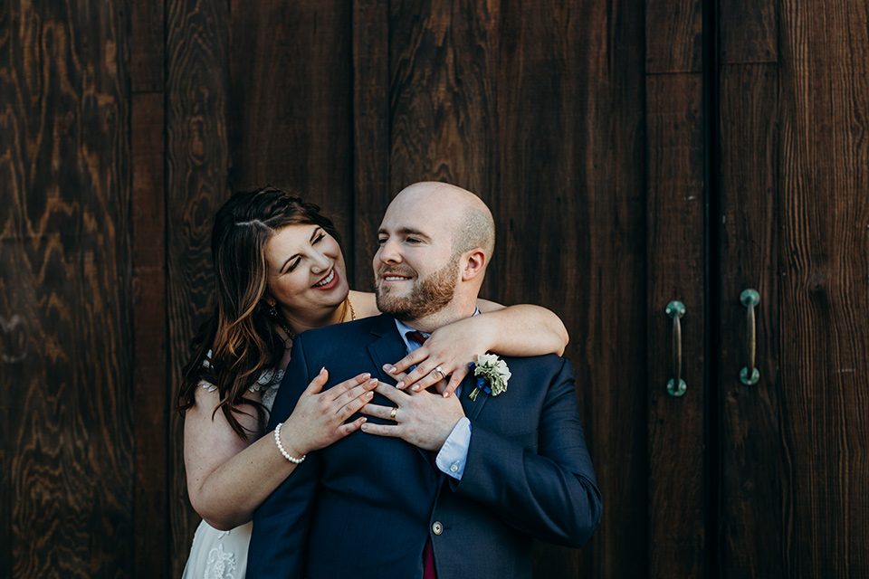 la-jolla-wedding-bride-and-groom-by-wooden-door-bride-in-a-flowing-ball-gown-with-a-detailed-bodice-and-straps-groom-in-a-dark-blue-suit-with-a-light-blue-suit-and-red-tie