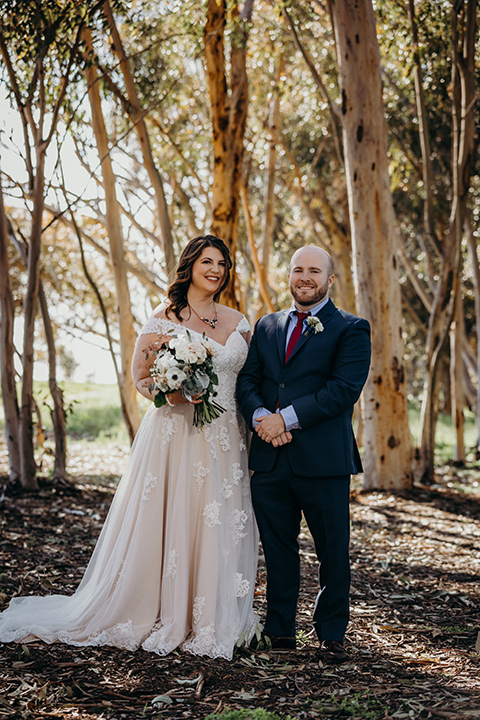 la-jolla-wedding-bride-and-groom-smiling-at-camera-by-trees-bride-in-a-tulle-ballgown-with-a-crystaled-bodice-and-cap-sleeves-groom-in-a-navy-suit-and-light-blue-shirt