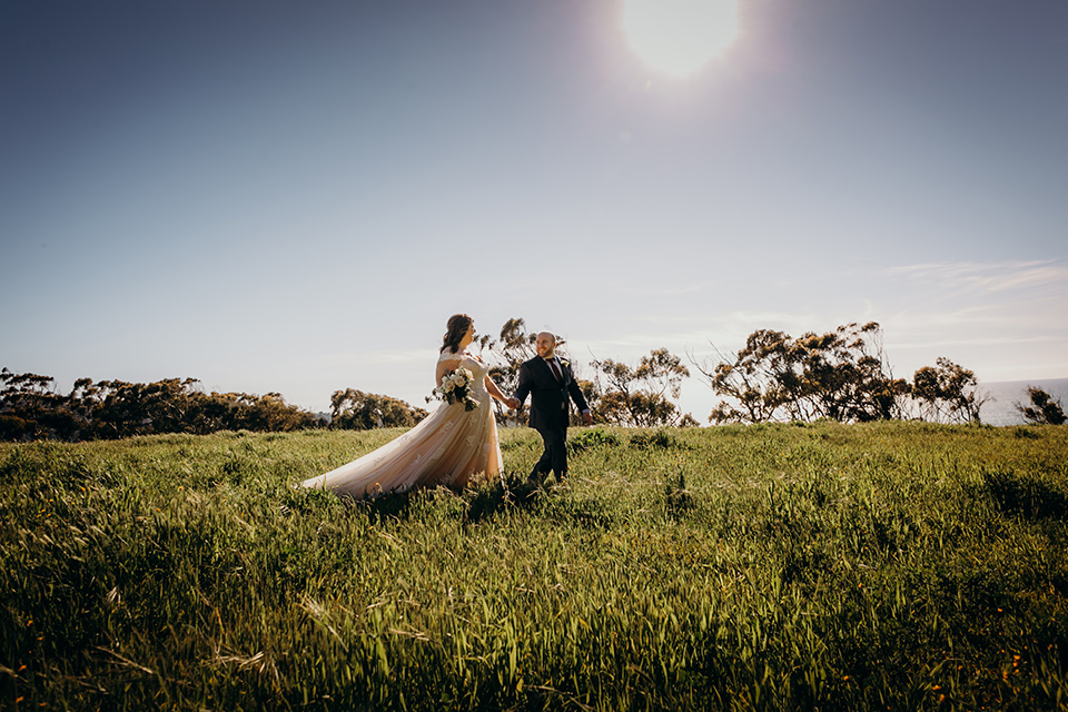 la-jolla-wedding-bride-and-groom-walking-in-grass-bride-in-a-flowing-ball-gown-with-a-detailed-bodice-and-straps-groom-in-a-dark-blue-suit-with-a-light-blue-suit-and-red-tie