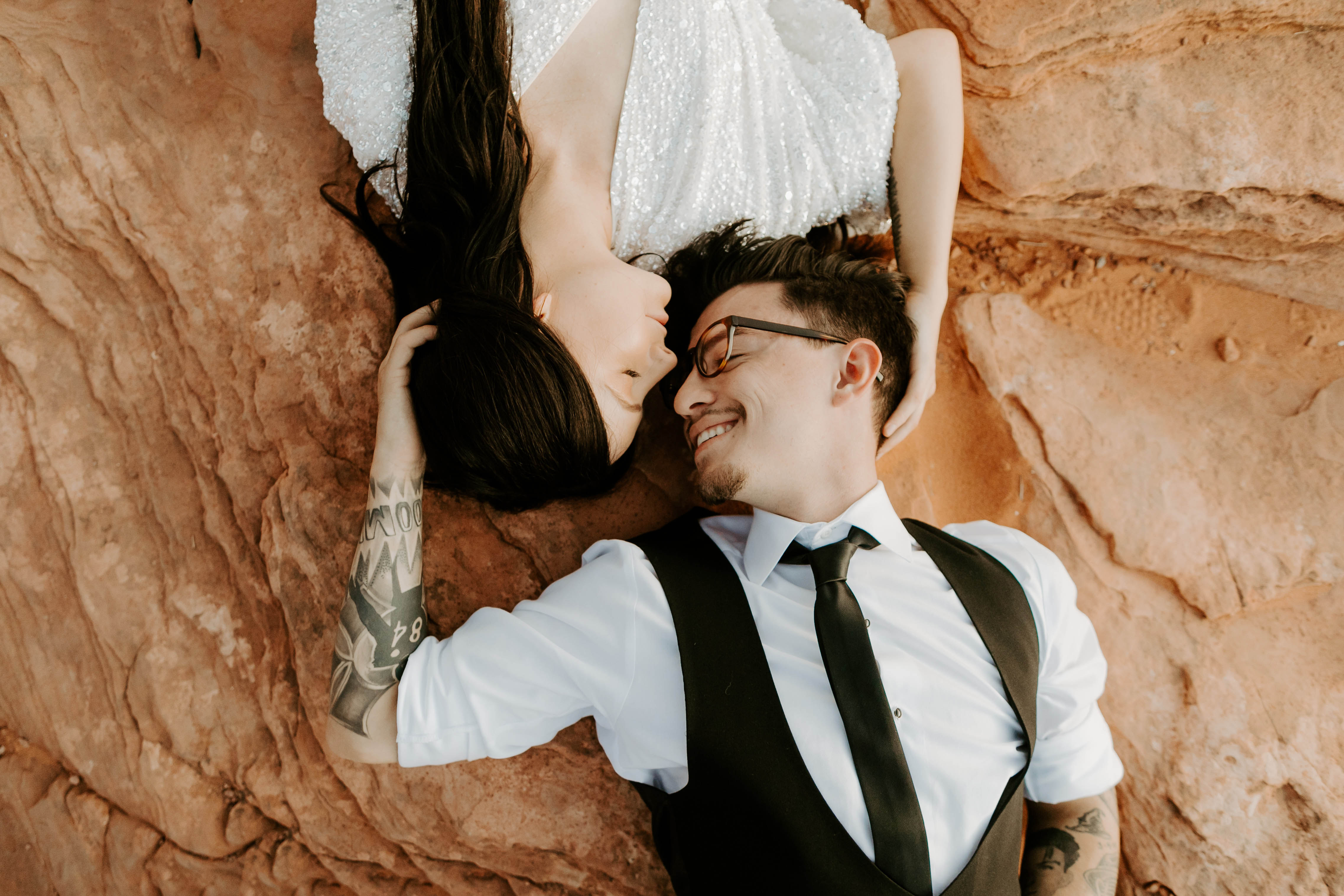  bride in an ivory long gown with bellowing sleeves and a boho design, groom in a black double-breasted vest and a white buttoned up shirt laying on the ground