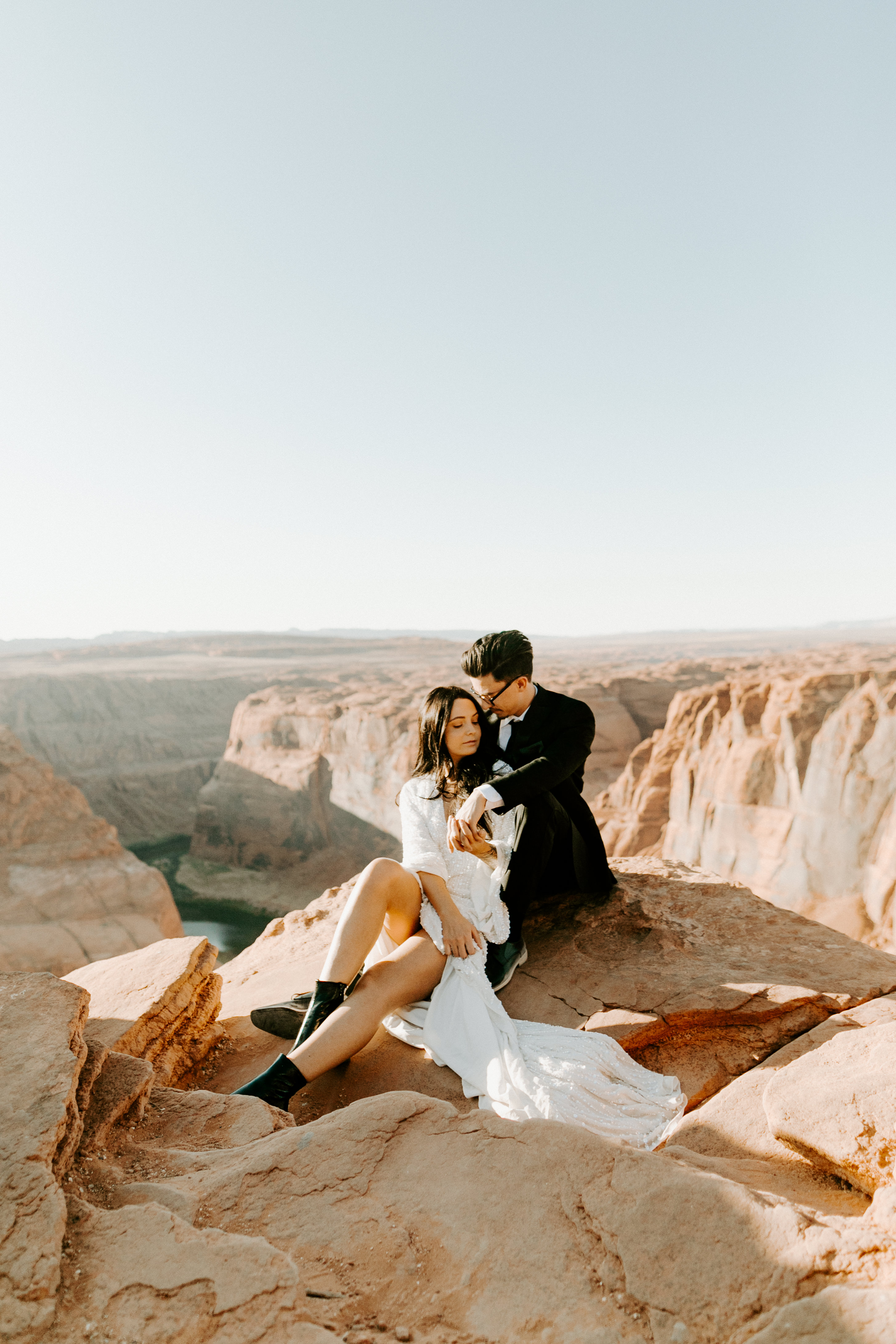  bride in an ivory long gown with bellowing sleeves and a boho design, groom in a black double-breasted vest and a white buttoned up shirt sitting on cliffside