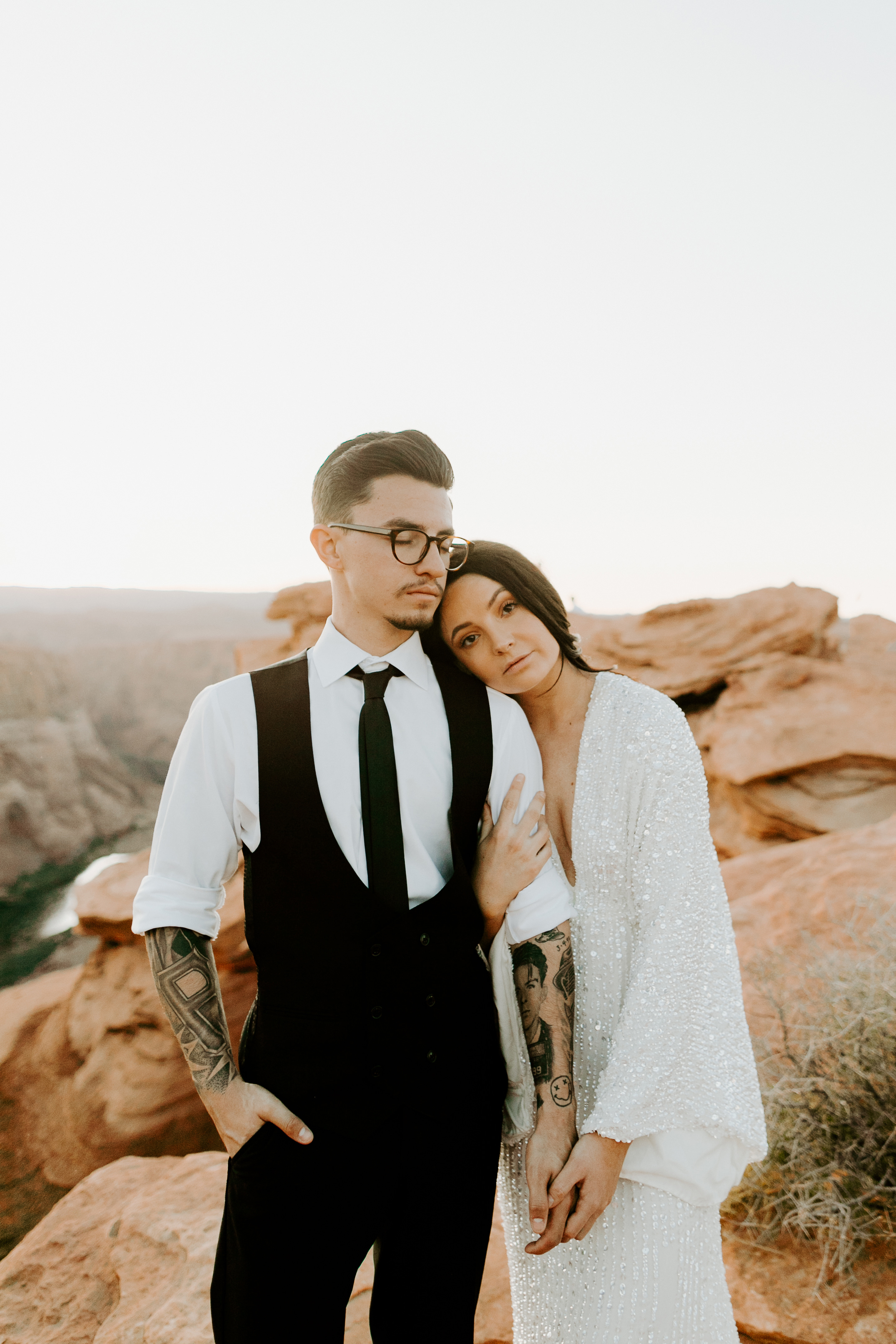  bride in an ivory long gown with bellowing sleeves and a boho design, groom in a black double-breasted vest and a white buttoned up shirt sitting on cliffside