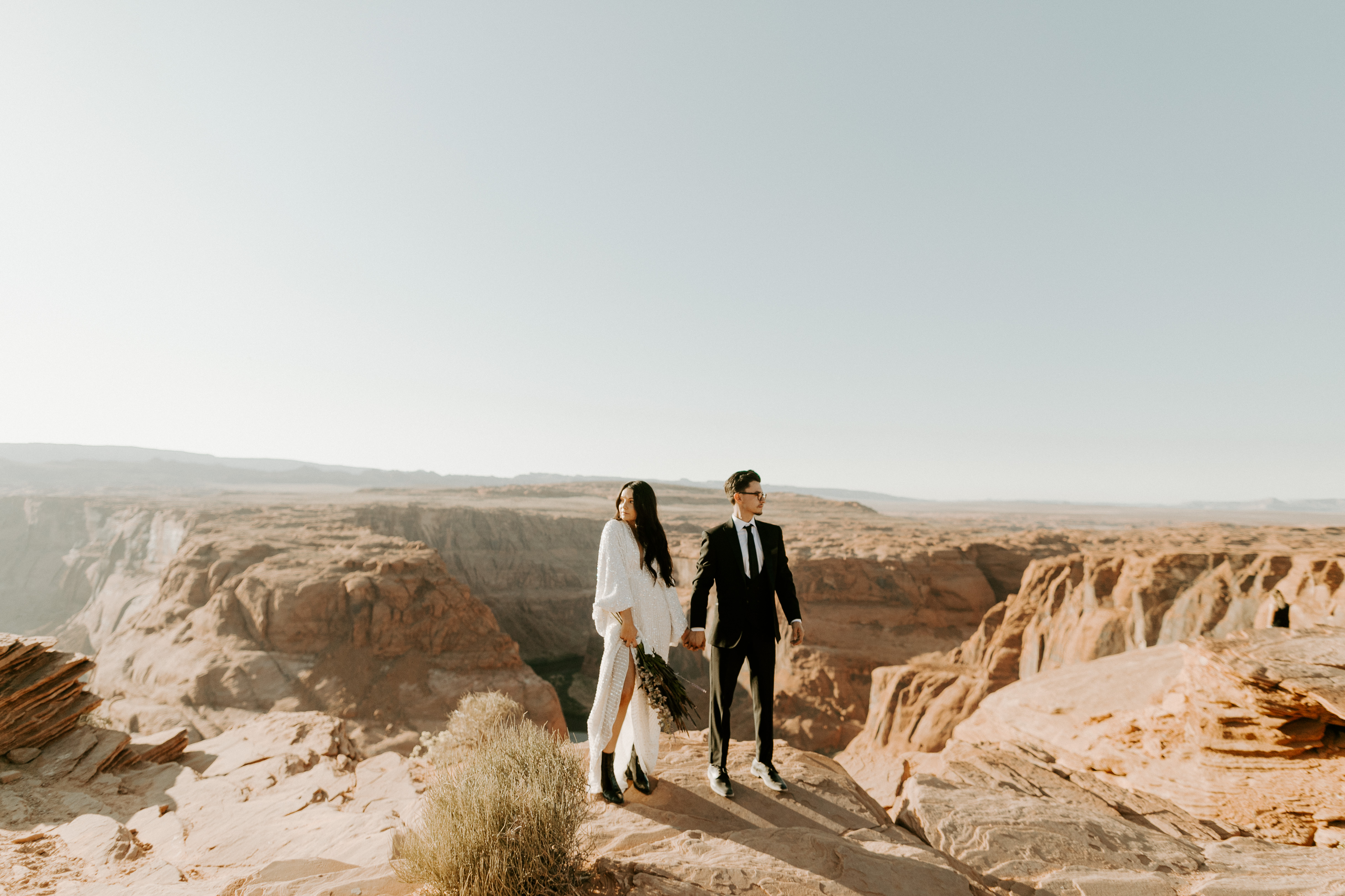  bride in an ivory long gown with bellowing sleeves and a boho design, groom in a black double-breasted vest and a white buttoned up shirt walking on horseshoe bend