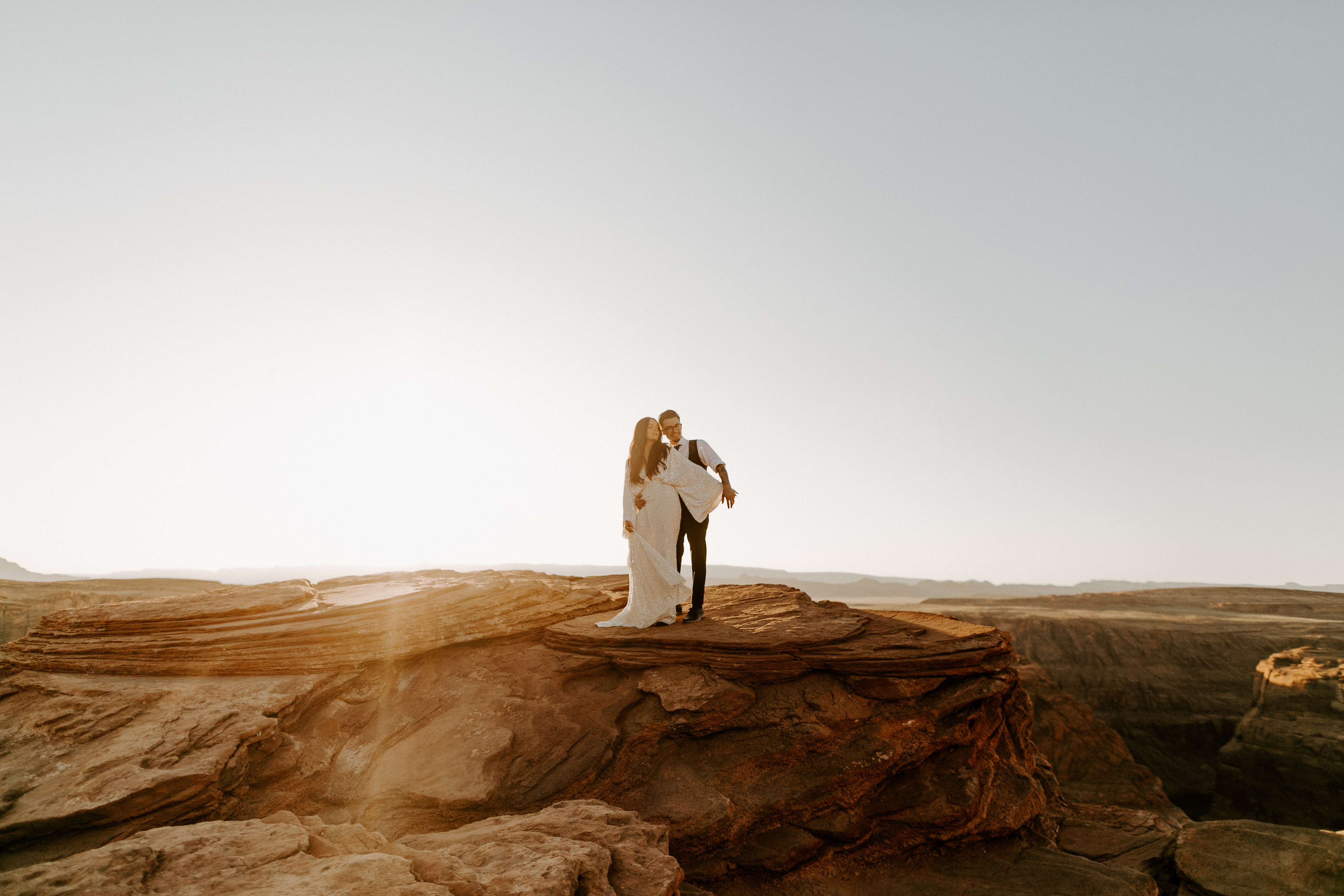  bride in an ivory long gown with bellowing sleeves and a boho design, groom in a black double-breasted vest and a white buttoned up shirt walking on horseshoe bend and cliffs