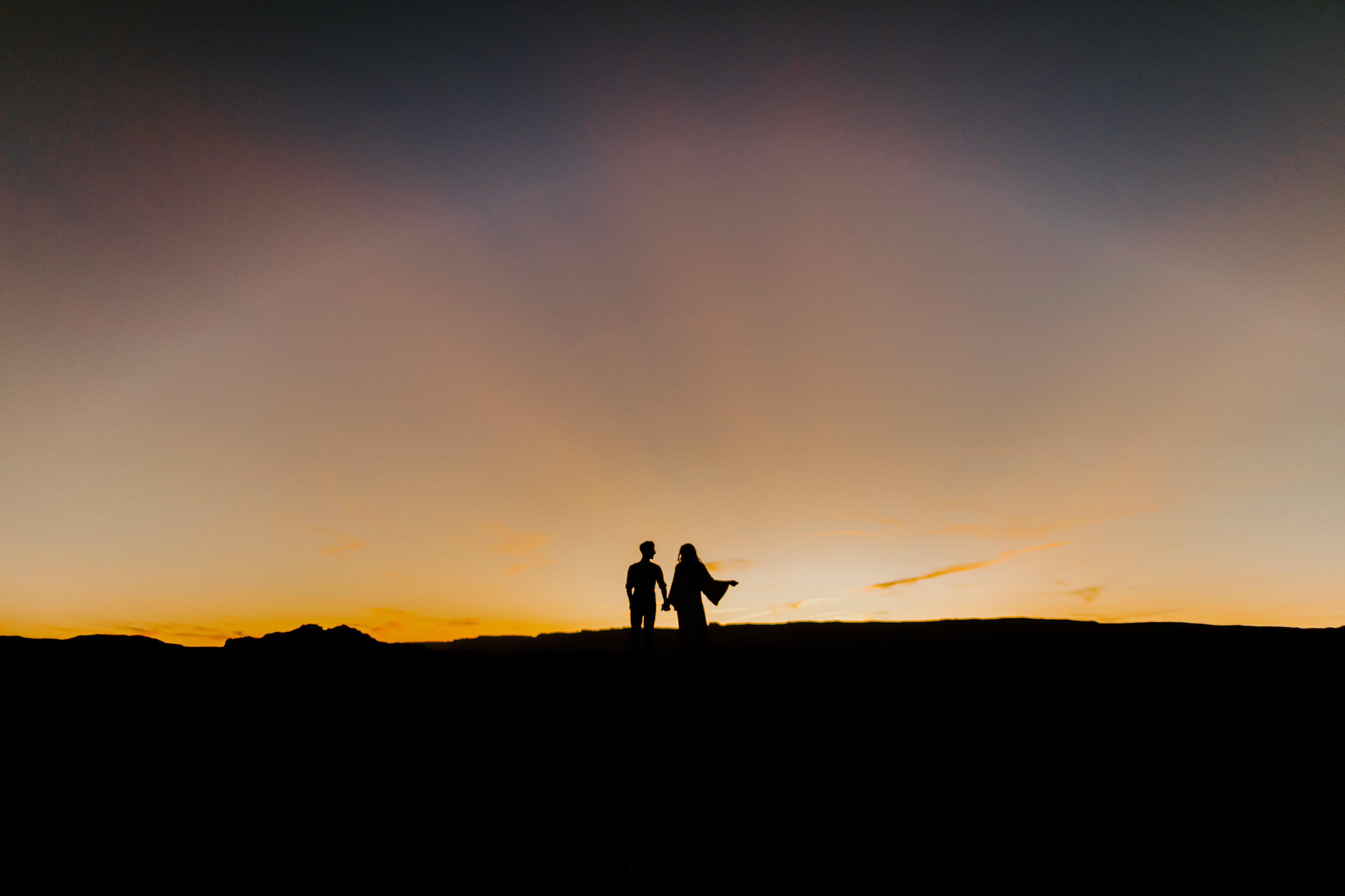  bride in an ivory long gown with bellowing sleeves and a boho design, groom in a black double-breasted vest and a white buttoned up shirt walking at sunset