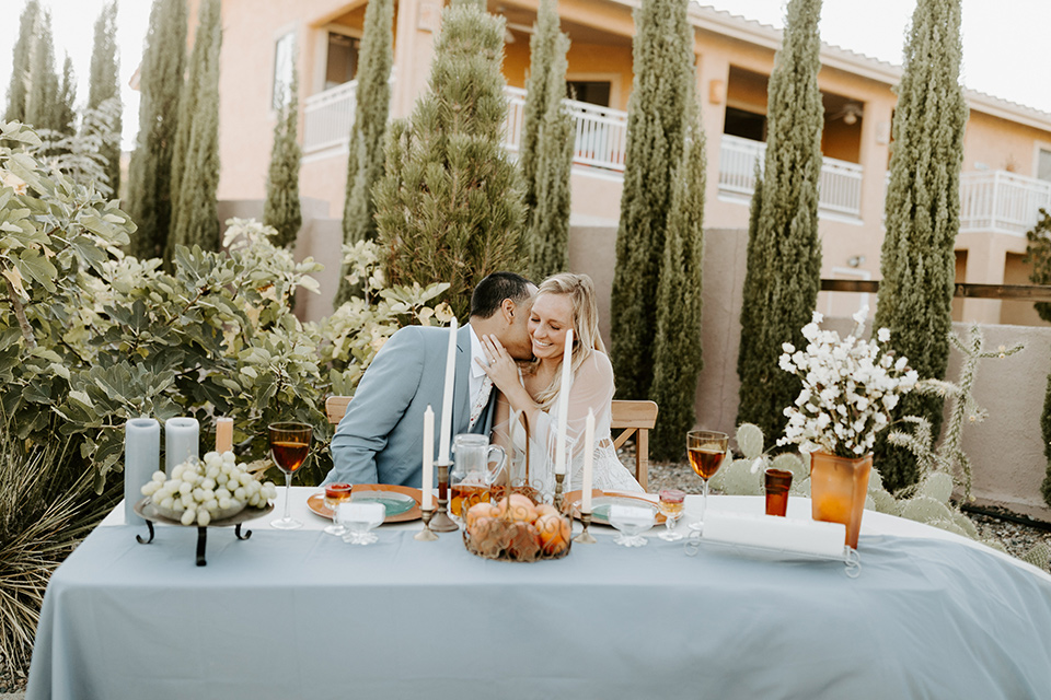  bride and groom sitting at sweetheart table, bride in an ivory lace gown with bellowing sleeves and a boho design.  The groom is in a light blue suit with matching vest and no tie