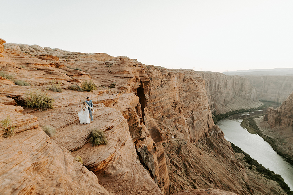  bride and groom standing on cliff in a faraway shot, bride in an ivory lace gown with bellowing sleeves and a boho design.  The groom is in a light blue suit with matching vest and no tie