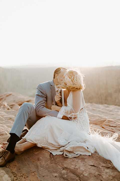  bride in an ivory lace gown with bellowing sleeves and a boho design, The groom is in a light blue suit with matching vest and no tie resting on the side of the cliff 