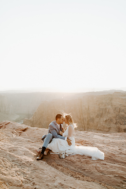  bride in an ivory lace gown with bellowing sleeves and a boho design, The groom is in a light blue suit with matching vest and no tie sitting on the side of the cliff