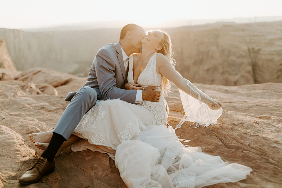  bride and groom standing on cliff in a faraway shot, bride in an ivory lace gown with bellowing sleeves and a boho design.  The groom is in a light blue suit with matching vest and no tie