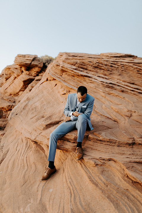  The groom is in a light blue suit with matching vest and no tie sitting on the side of the cliff