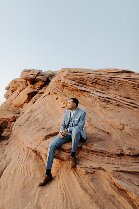  The groom is in a light blue suit with matching vest and no tie resting on the side of the cliff 