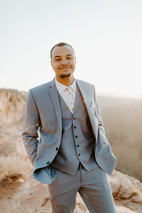  The groom is in a light blue suit with matching vest and no tie sitting on the side of the cliff