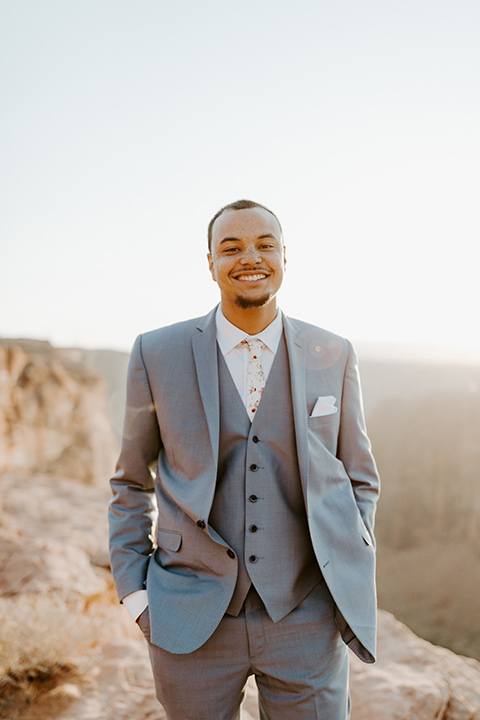  The groom is in a light blue suit with matching vest and no tie resting on the side of the cliff 