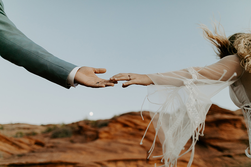  bride and groom standing on cliff in a faraway shot, bride in an ivory lace gown with bellowing sleeves and a boho design.  The groom is in a light blue suit with matching vest and no tie