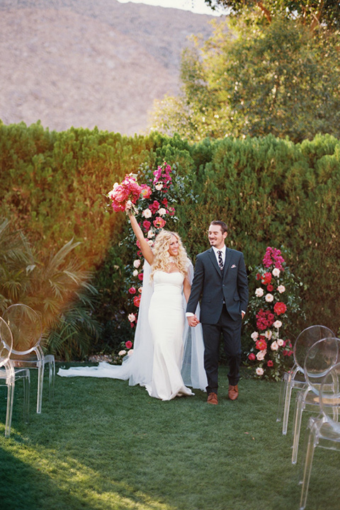  bride in a vintage bohemian gown with beaded details and cape, groom in a green suit with a floral tie at ceremony