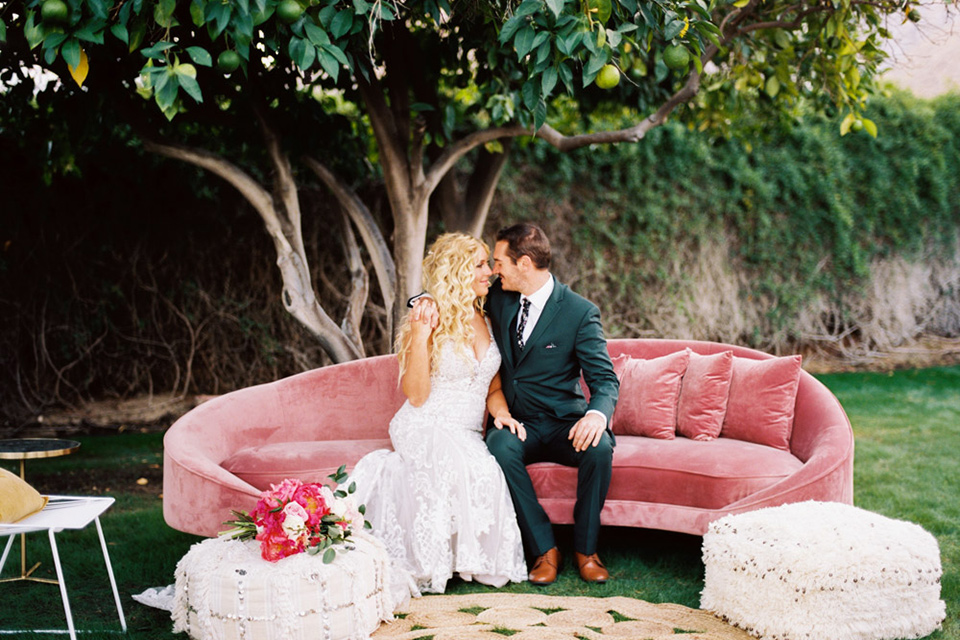  bride in a vintage bohemian gown with beaded details and cape, groom in a green suit with a floral tie sitting on couch
