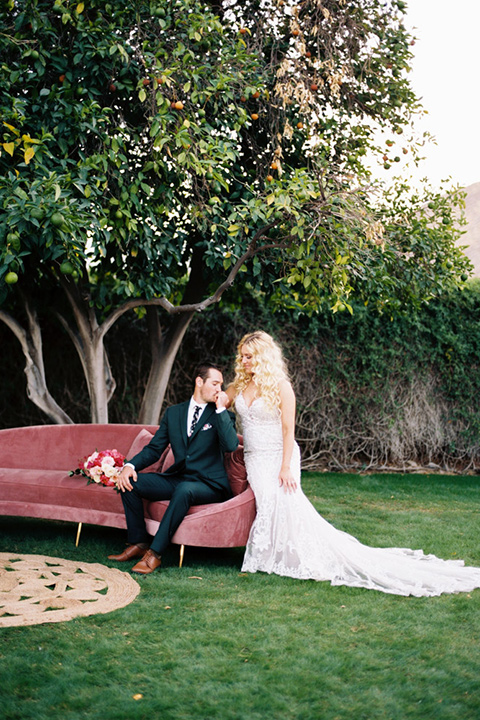  bride in a vintage bohemian gown with beaded details and cape, groom in a green suit with a floral tie sitting on couch