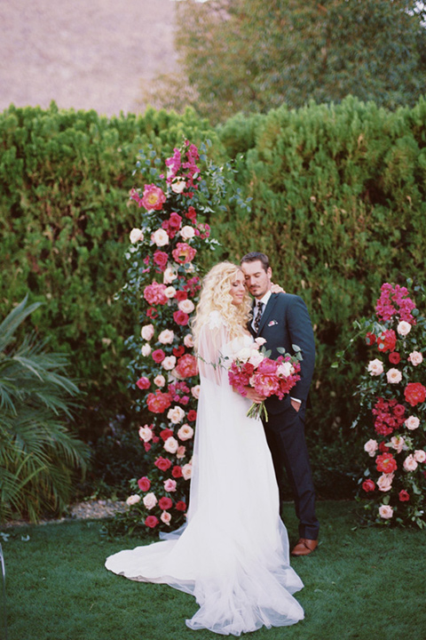  bride in a vintage bohemian gown with beaded details and cape, groom in a green suit with a floral tie at ceremony