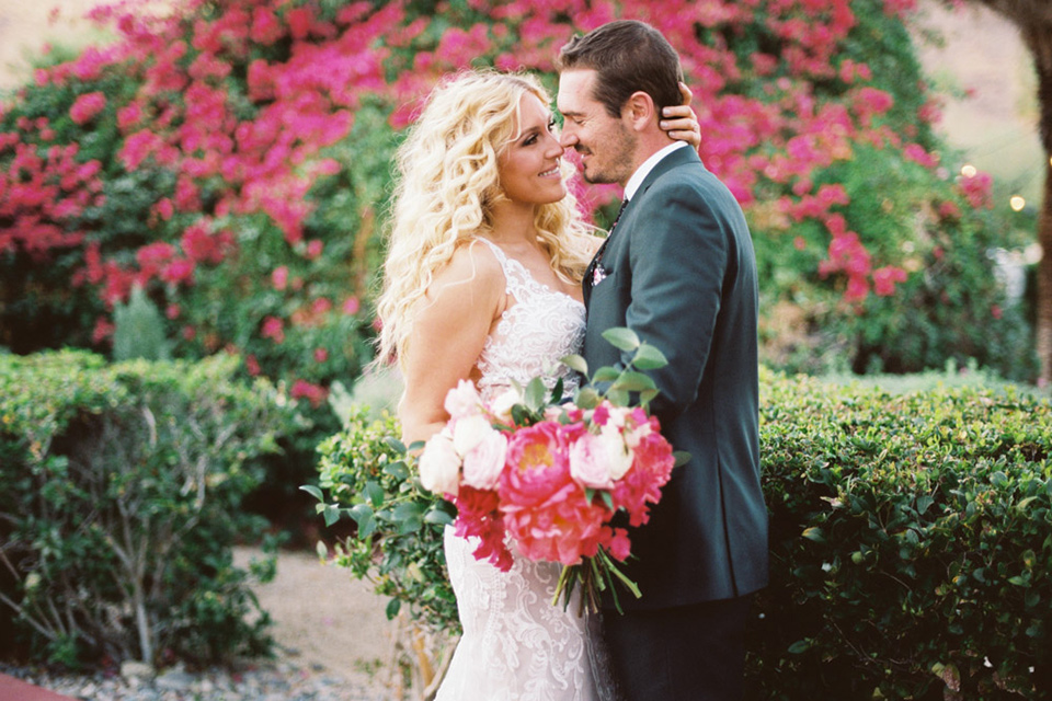  bride in a vintage bohemian gown with beaded details and cape, groom in a green suit with a floral tie sitting on couch